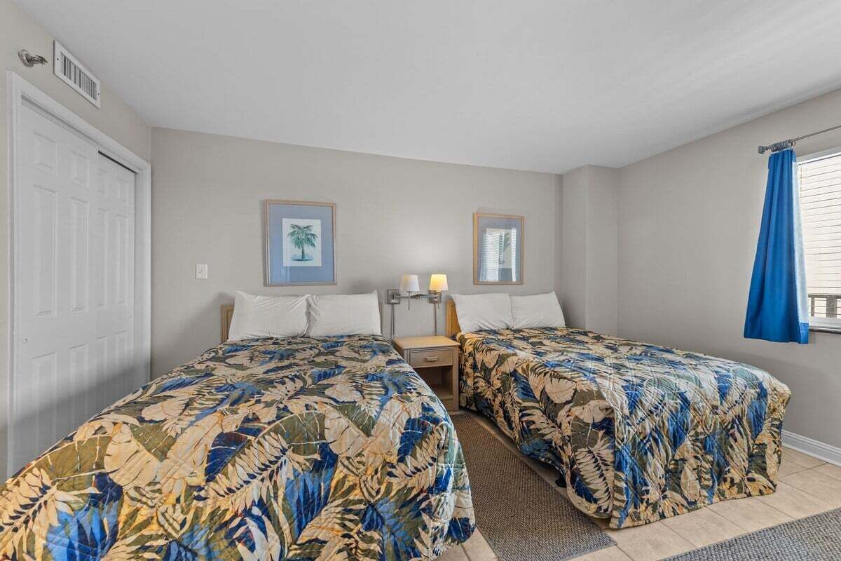A room with two beds, each with a floral bedspread, a nightstand with a lamp, and a window with blue curtains.