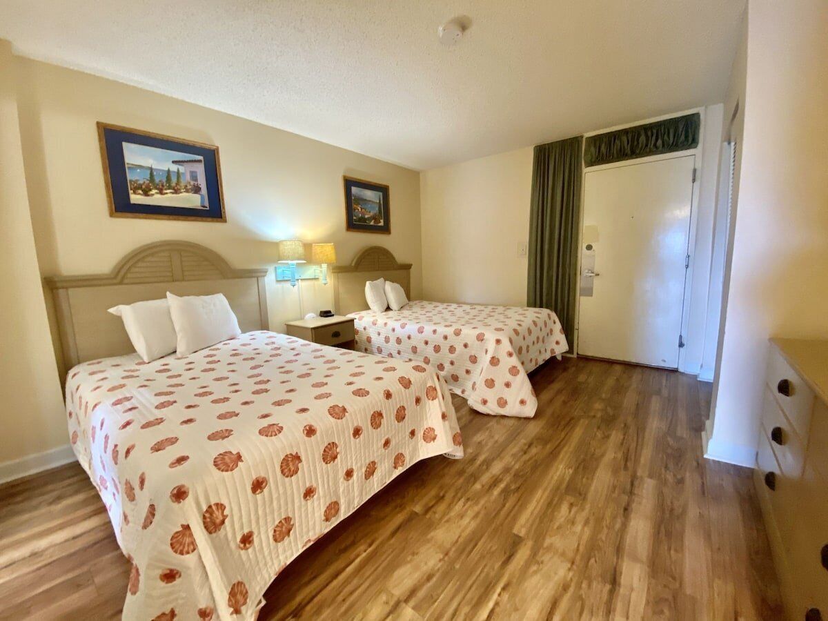 A hotel room with two beds, wooden flooring, a nightstand with a lamp, and a door with a green curtain.