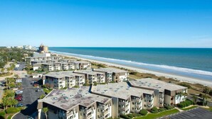 A coastal hotel with multiple-story buildings, a parking lot, and a view of the beach and ocean.
