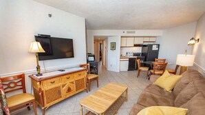 A living room with a sofa, wooden coffee table, and a television. There is a kitchen area with cabinets and a refrigerator.