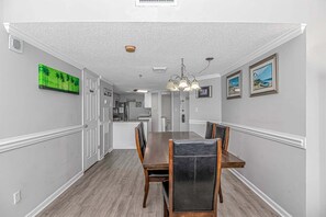 A dining area with a long table, chairs, a chandelier, and kitchen in the background.