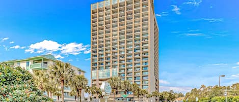 A tall hotel with multiple floors, a green building to the left, and palm trees in the foreground.