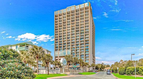 A tall hotel with multiple floors, a green building to the left, and palm trees in the foreground.