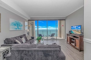 Uma sala de estar com um sofá cinzento, uma mesa de centro com uma planta, uma televisão sobre um suporte de madeira e uma varanda com vista para o mar.