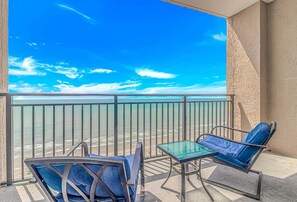 A balcony with a clear view of the beach and ocean. There are two blue chairs and a small table.