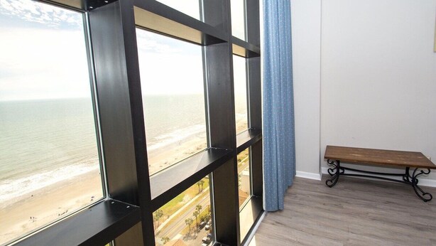 Una habitación con ventanas amplias que dan a una playa, un banco de madera y un suelo de color claro.
