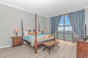 A four-poster bed with a bench, a nightstand, and a television in a room with a large window and a view of the sea.