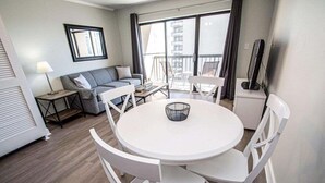 A modern living room with a round white table, white chairs, a grey sofa, and a balcony with a view of buildings. - 1Br/1Ba MB Condo! Sleeps 6! Steps To The Ocean! (Myrtle Beach)