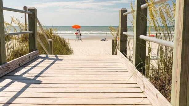 A wooden boardwalk leading to a beach with a person holding an umbrella. - 1Br/1Ba MB Condo! Sleeps 6! Steps To The Ocean! (Myrtle Beach)