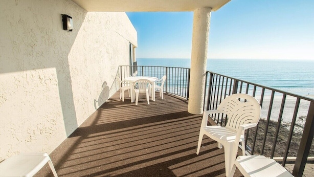 A balcony with a view of the sea, featuring a table and chairs, a railing, and a textured wall.
