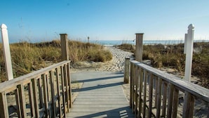 A wooden boardwalk leading to a beach with sand and grass on either side.