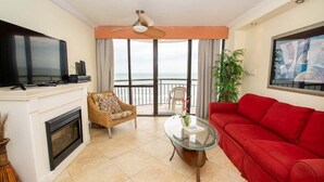 Una sala de estar con televisor de pantalla plana, un sofá rojo, una mesa de centro de vidrio, un sillón de mimbre y vistas al océano a través de amplios ventanales.