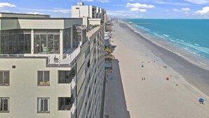 Un hôtel en bord de mer, avec plusieurs étages, des balcons et une plage de sable, avec une jetée en arrière-plan.