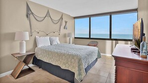 Une chambre à coucher comprenant un lit, deux tables de chevet, une commode, une coiffeuse et une grande fenêtre donnant sur l’océan.