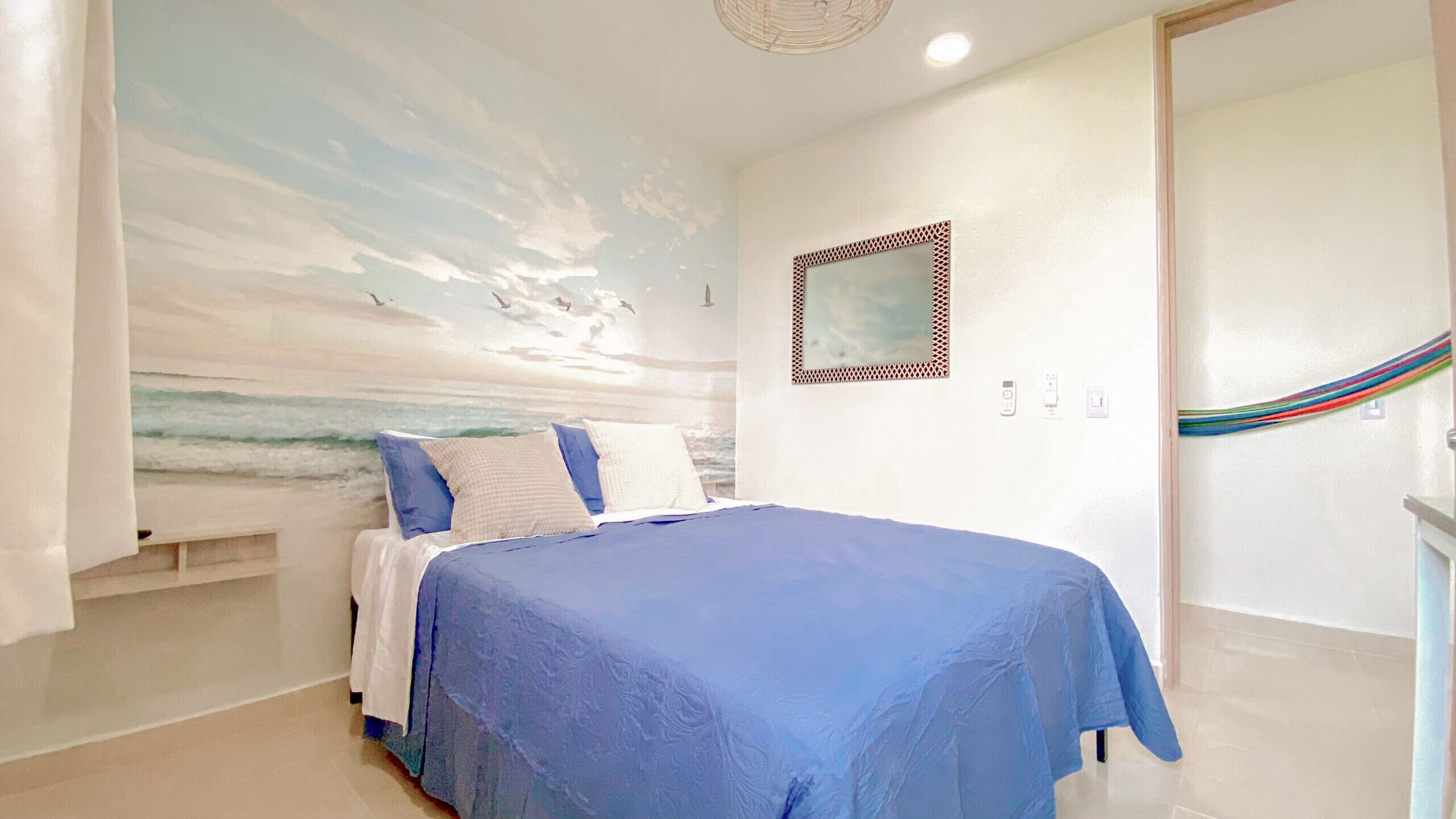 Ein Schlafzimmer mit einem Bett, einer Wandmalerei mit Strandmotiv, einer Tür mit bunter Vorhang und einem kleinen Regal.