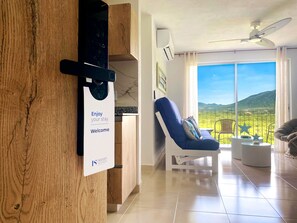 Quarto de hotel moderno com uma mesa de recepção de madeira, uma poltrona reclinável azul, uma varanda com vista para o campo e um ventilador de teto.