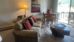 A living room with a sofa, a table with chairs, a lamp, and a large window. - Book now for great fall colors…watch wildlife from deck! (Angel Fire)