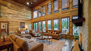 A spacious living room with multiple sofas and a fireplace, large windows offering views of the outdoors, and a wooden ceiling. - The Unicorn - Featured on Staycation: Luxury Broken Bow Retreat (Broken Bow)