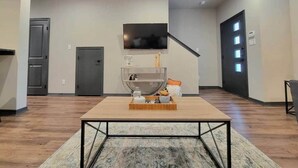 A spacious room with a wooden floor, a central table with a tray of items, a mounted TV, and a door with a light switch.
