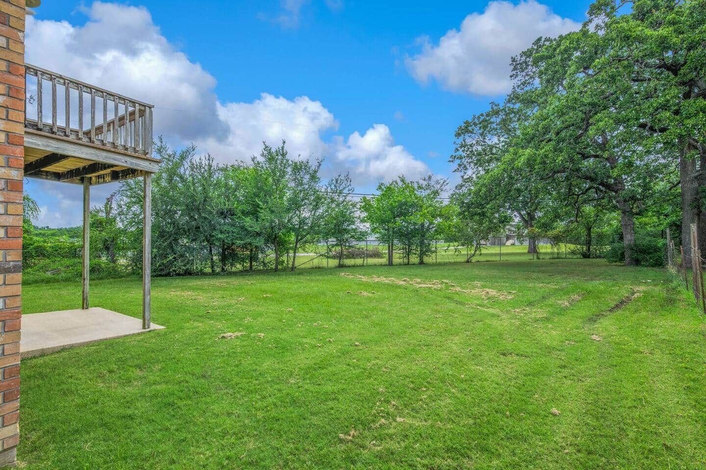 A backyard with a wooden deck, a brick wall, and a grassy lawn with trees.