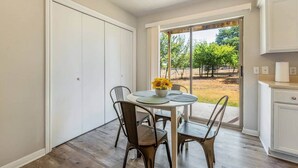 Un comedor con mesa y sillas, vista a un jardín a través de una puerta corrediza de vidrio, y una zona de cocina con armarios.