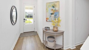 Un couloir avec une porte blanche, un miroir, un tableau et une console sur laquelle est posé un vase de fleurs.