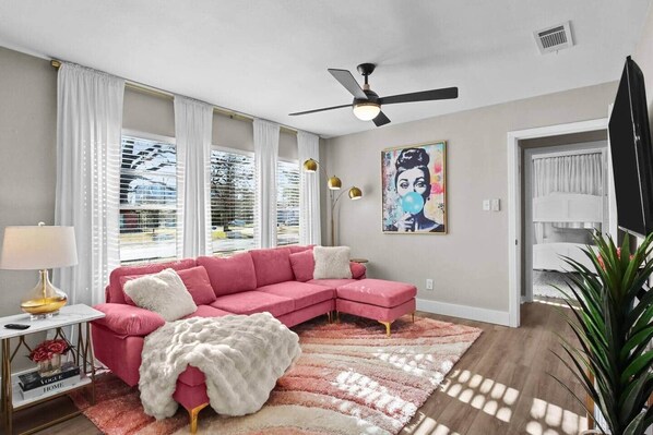 A modern living room with a pink sectional sofa, a ceiling fan, and a large window with blinds. - Chic Retreat • Stylish Stay near A&M (Bryan)