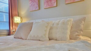 Un lit bien fait, avec une couette blanche et quatre oreillers, une lampe de chevet et une œuvre d’art abstraite rose accrochée au mur.