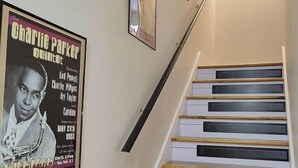 A staircase with wooden steps and white walls, a framed Charlie Parker poster, and a Jazz Houzz logo. - Jazz Houzz @ Aggieland (College Station)