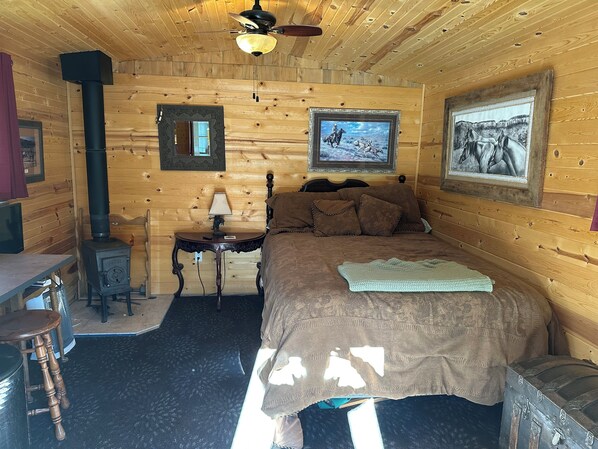 Ein Schlafzimmer einer Holzhütte mit einem Bett, einem kleinen Tisch mit Lampe, einem Heizgerät und gerahmten Bildern an den Wänden.