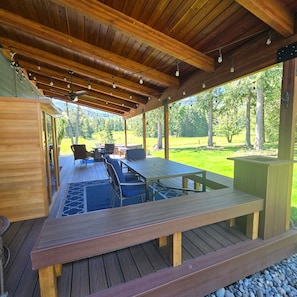 A covered outdoor area with a wooden deck, a table, chairs, and a view of a grassy landscape. - ElkLandia- A spacious 5+bd Home in Sunny Sequim, WA -Horses & Dogs welcome! (Sequim)