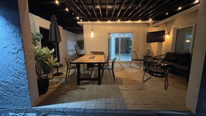 Un patio al aire libre con mesa y sillas de madera, un sofá y un televisor montado en la pared.