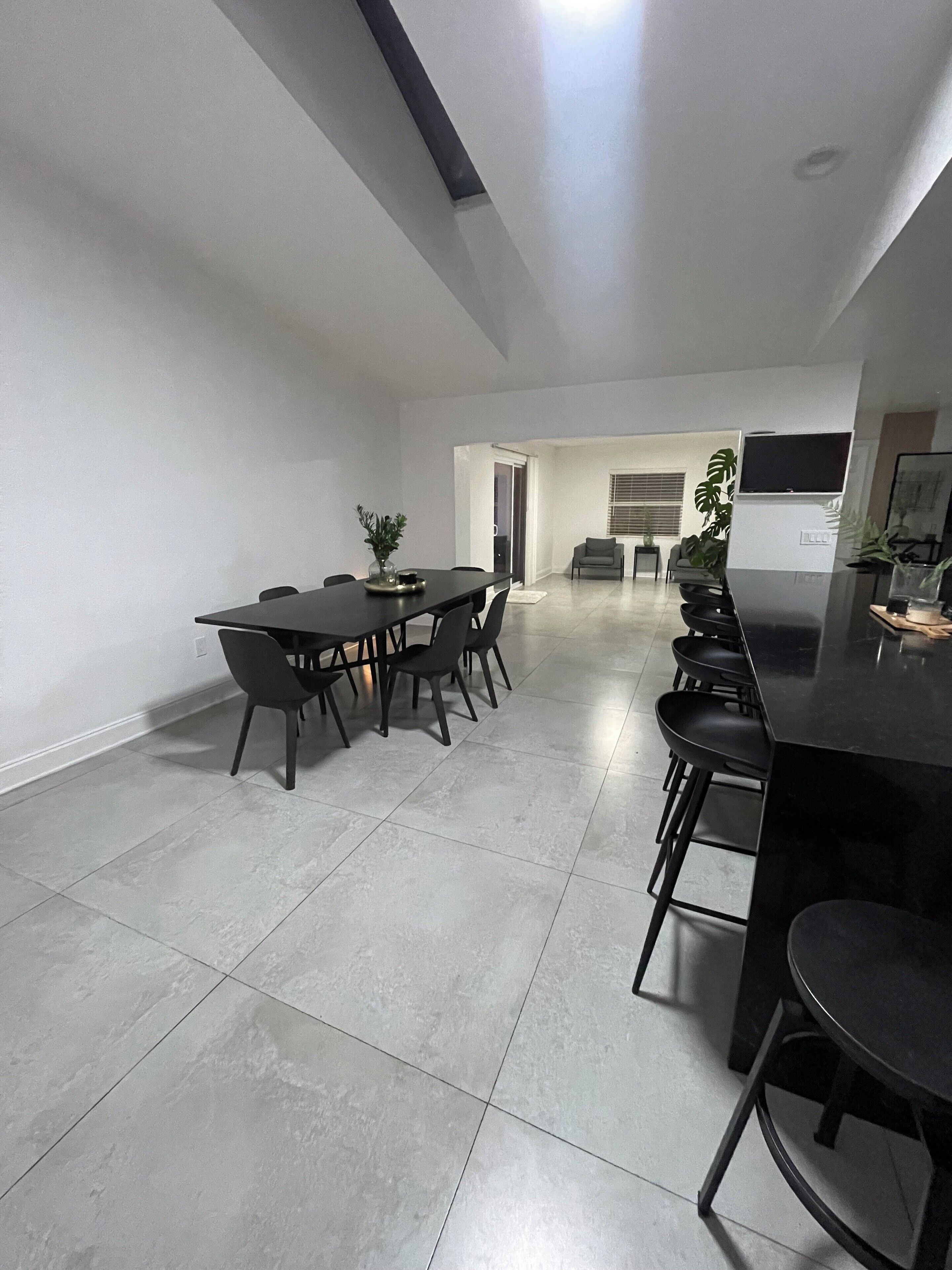 A modern kitchen with a dining area, featuring a long table and bar stools, and a small seating area with a TV.
