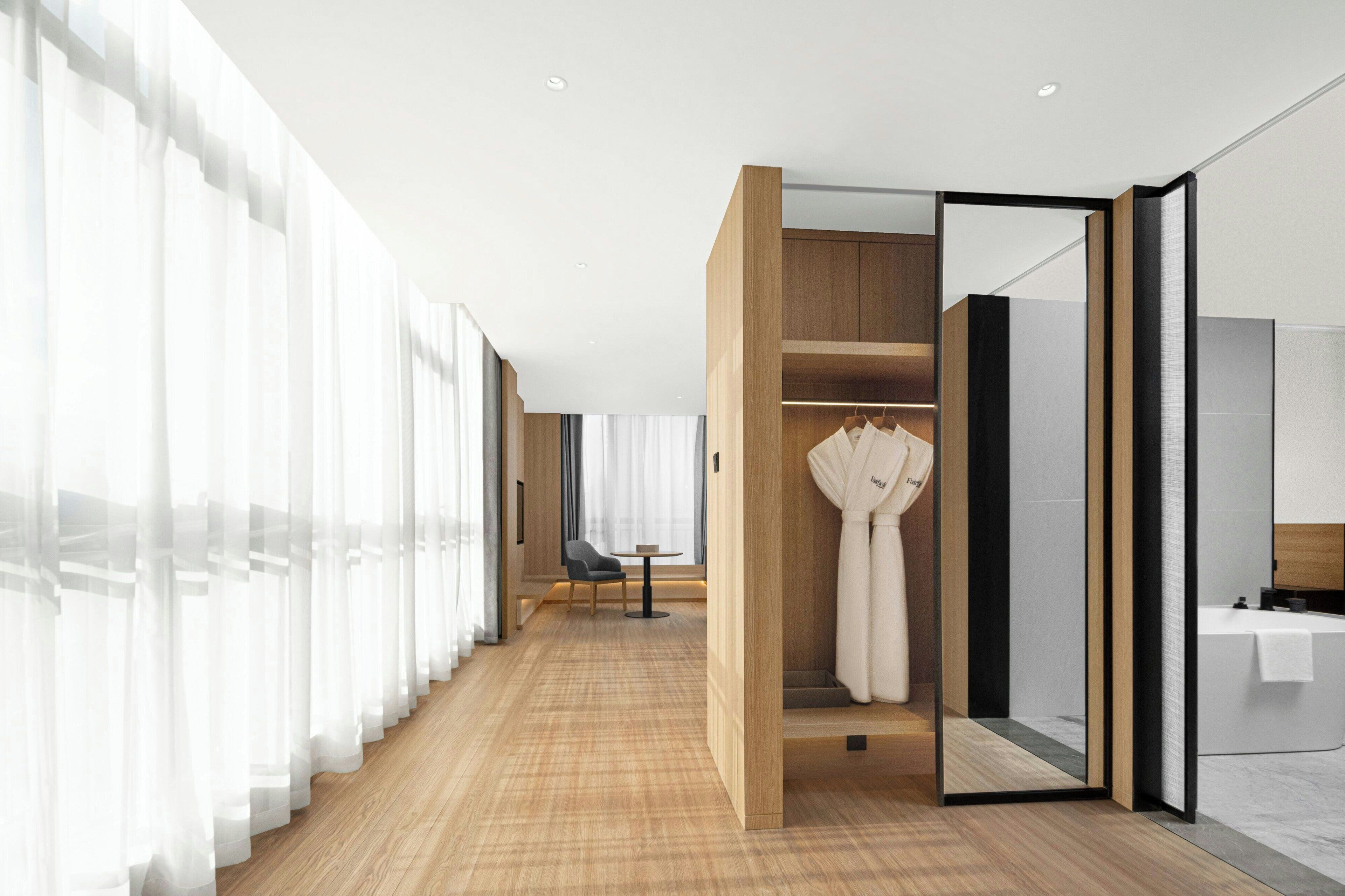 Een hotelkamer met een houten vloer, een zithoek met een stoel en een tafel, en een inloopkast met aanrechtjassen.