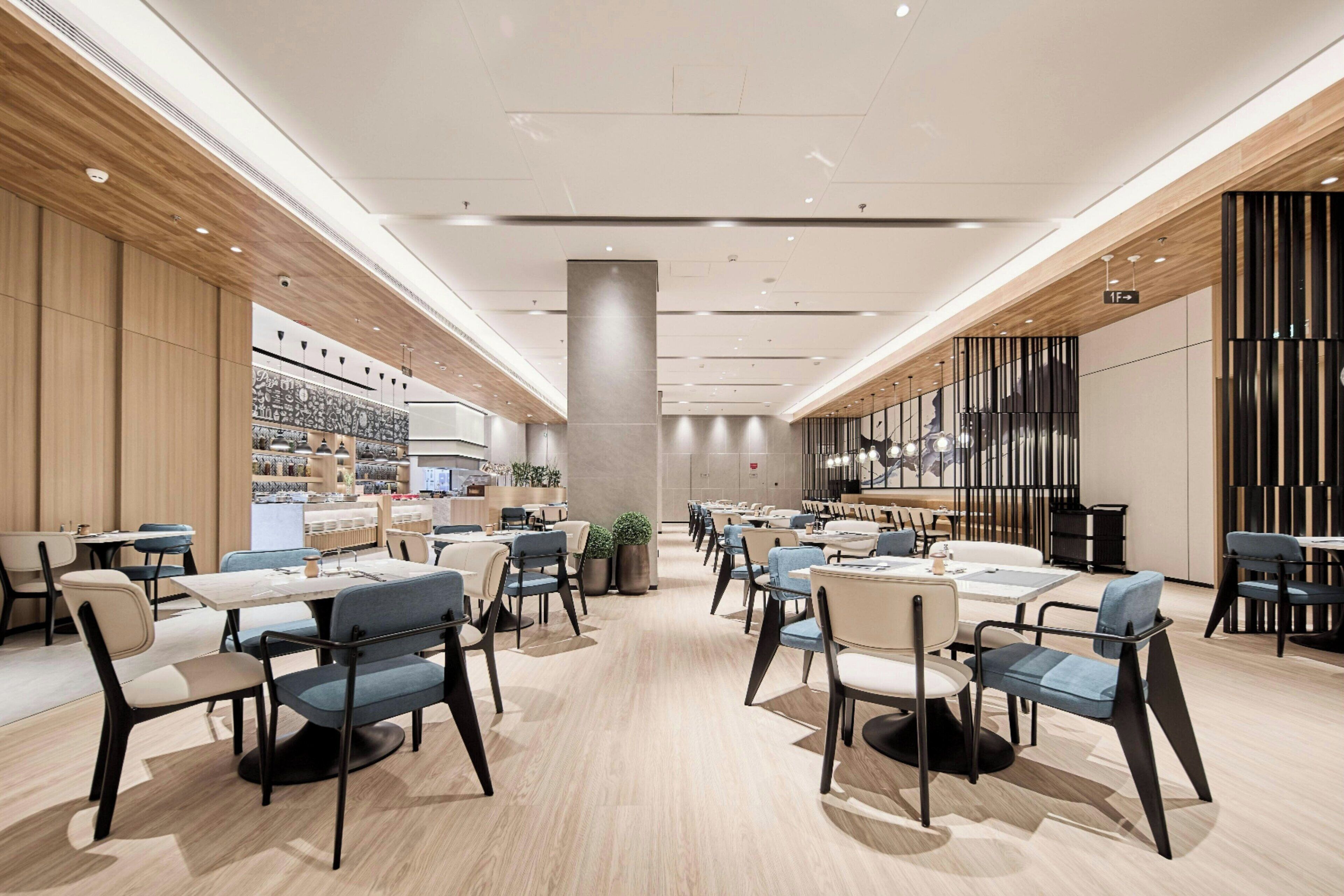 A spacious, modern dining area with multiple tables and chairs, a central pillar, and a kitchen area in the background.
