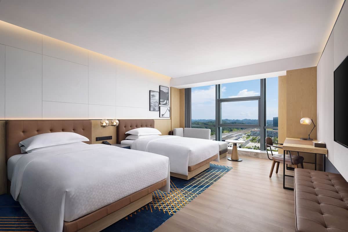 Ein modernes Hotelzimmer mit zwei Betten, einem Schreibtisch, einem Stuhl und einem großen Fenster mit Blick auf die Stadt.
