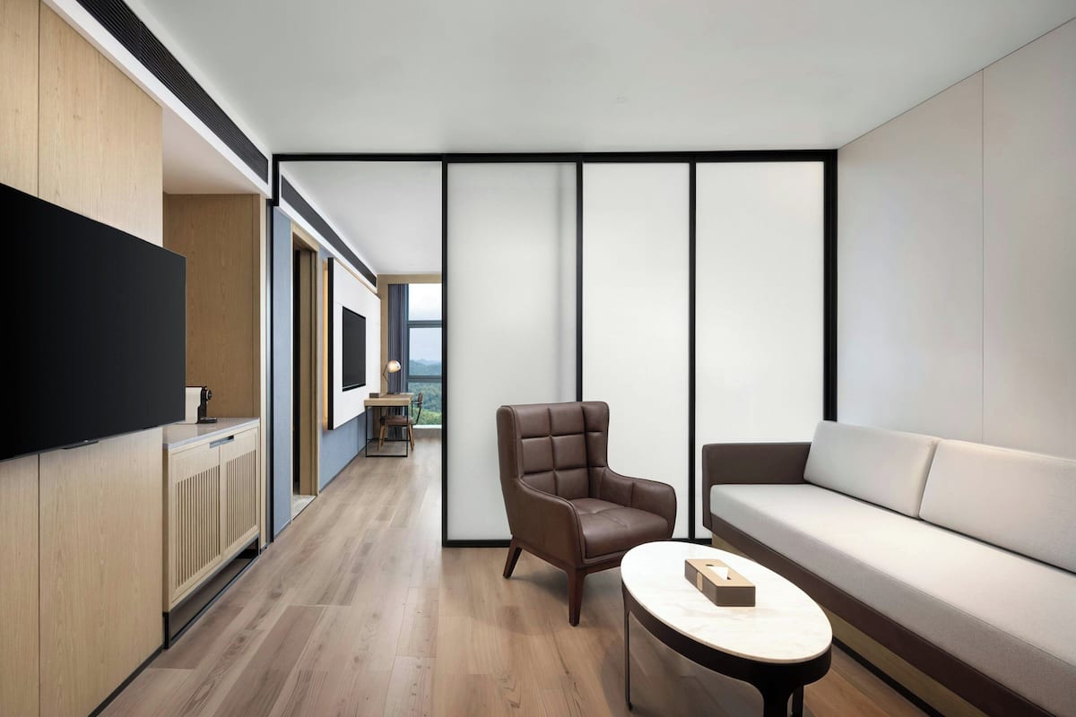 Ein modernes Wohnzimmer mit einer Couch, einem Ledersessel und einem Couchtisch. Links befindet sich ein Fernseher und durch ein großes Fenster bietet sich ein Blick ins Freie.