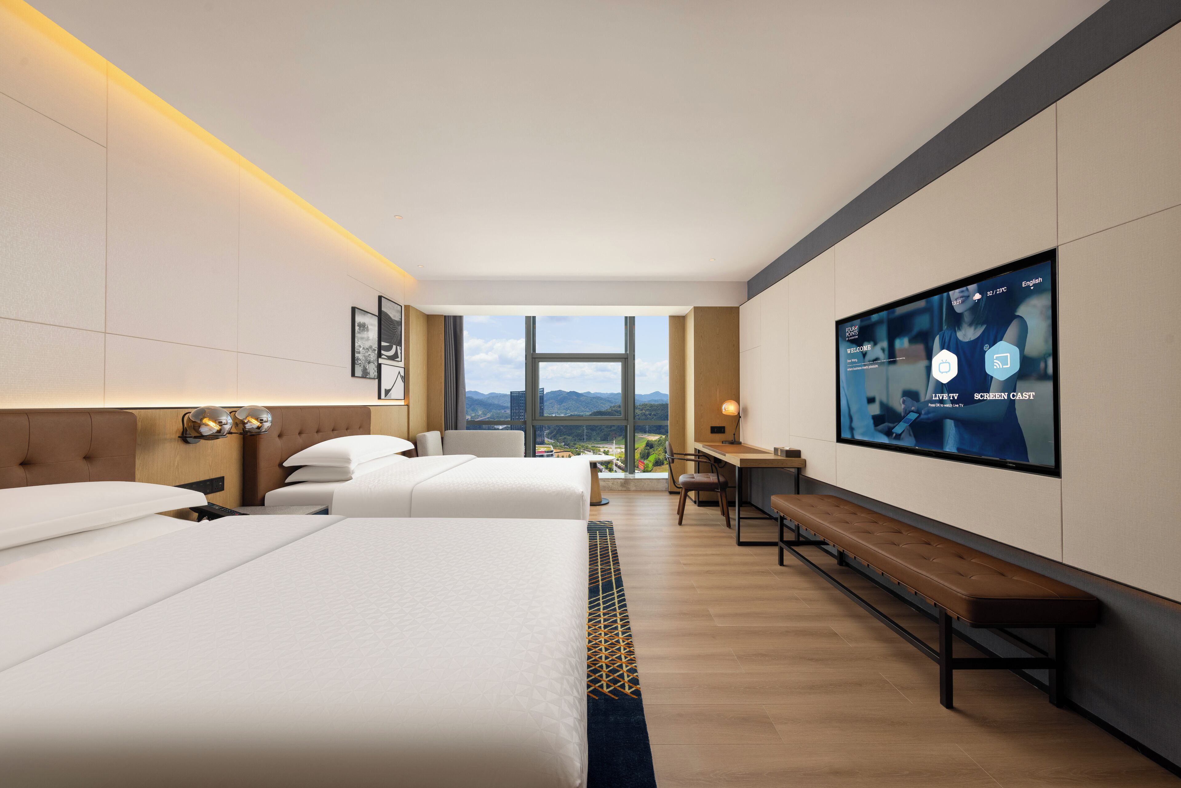 A hotel room with two beds, a desk, a TV, and a view of the city.