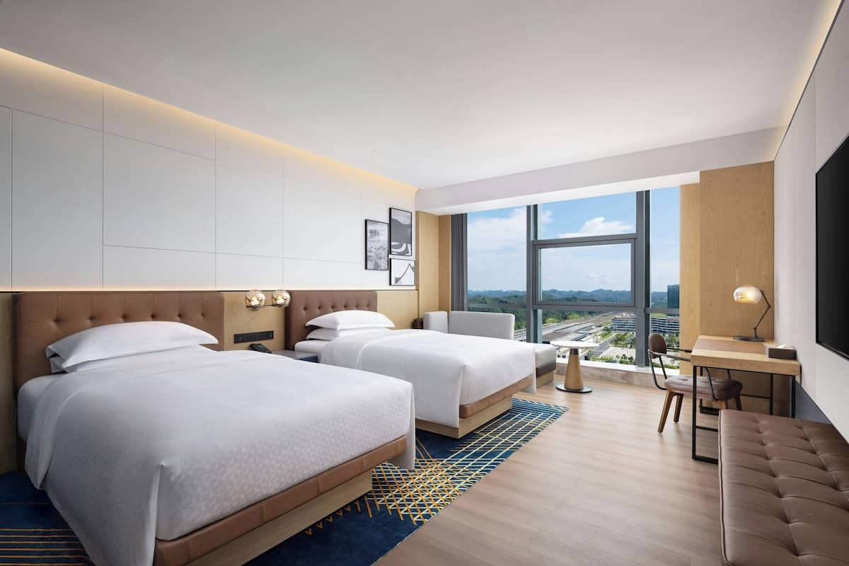 Ein modernes Hotelzimmer mit zwei Betten, einem Schreibtisch, einem Stuhl und einem großen Fenster mit Blick auf die Stadt.