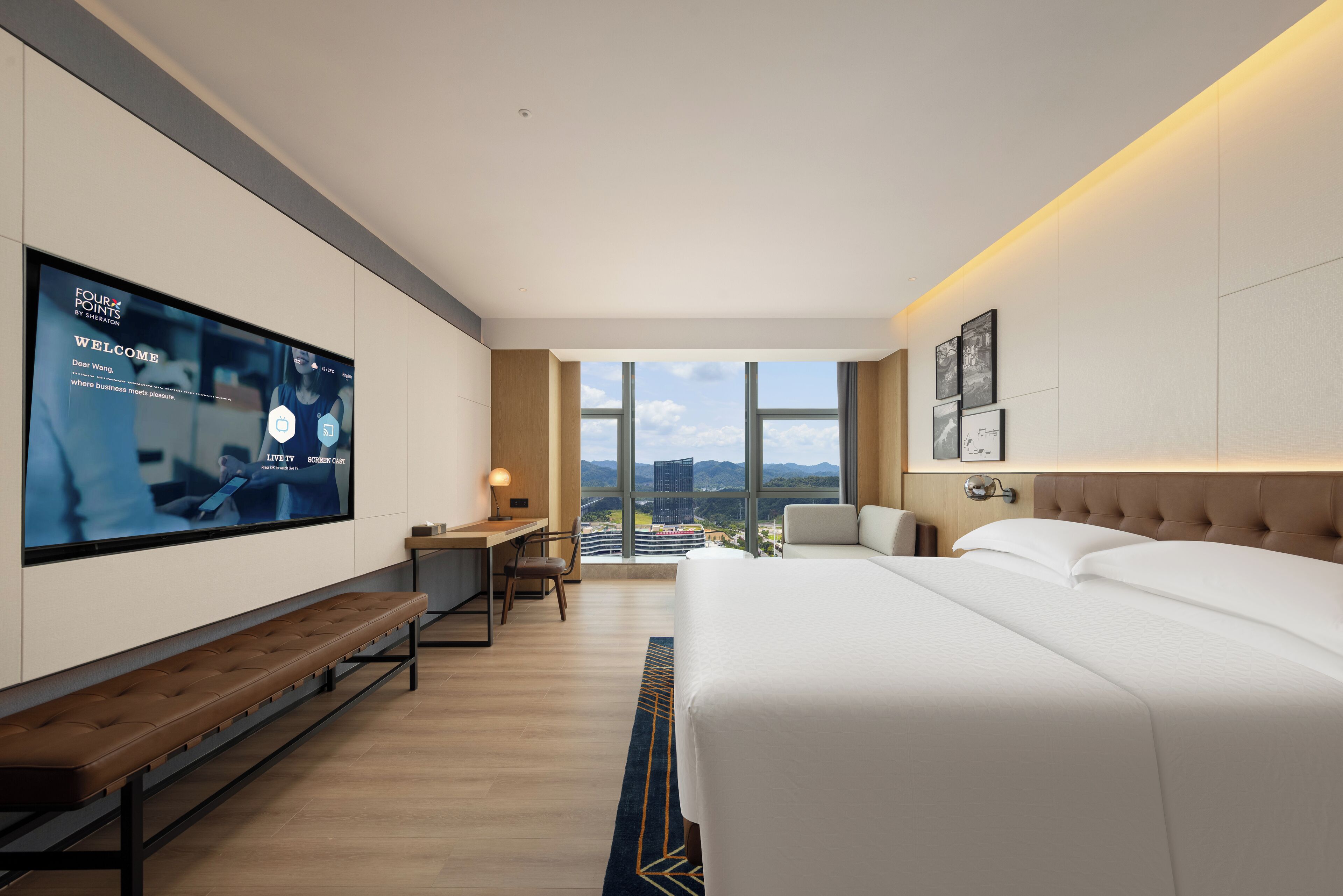 Habitación de hotel moderna con una cama grande, un escritorio con computadora, un televisor montado en la pared y vistas a la ciudad a través de amplias ventanas.