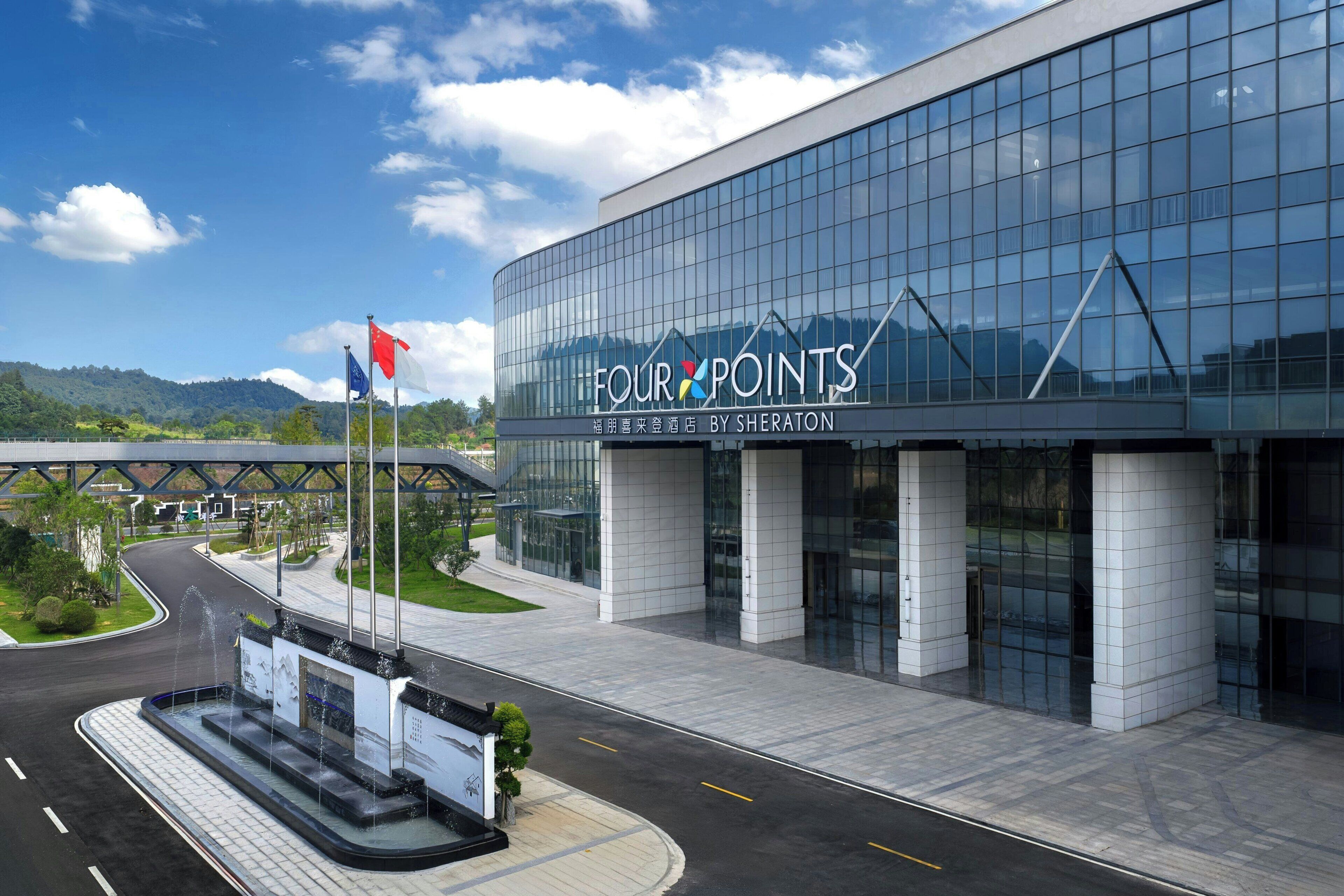 A modern hotel with a glass facade, a sign reading "FOUR POINTS BY SHERATON", and a landscaped area with a fountain.