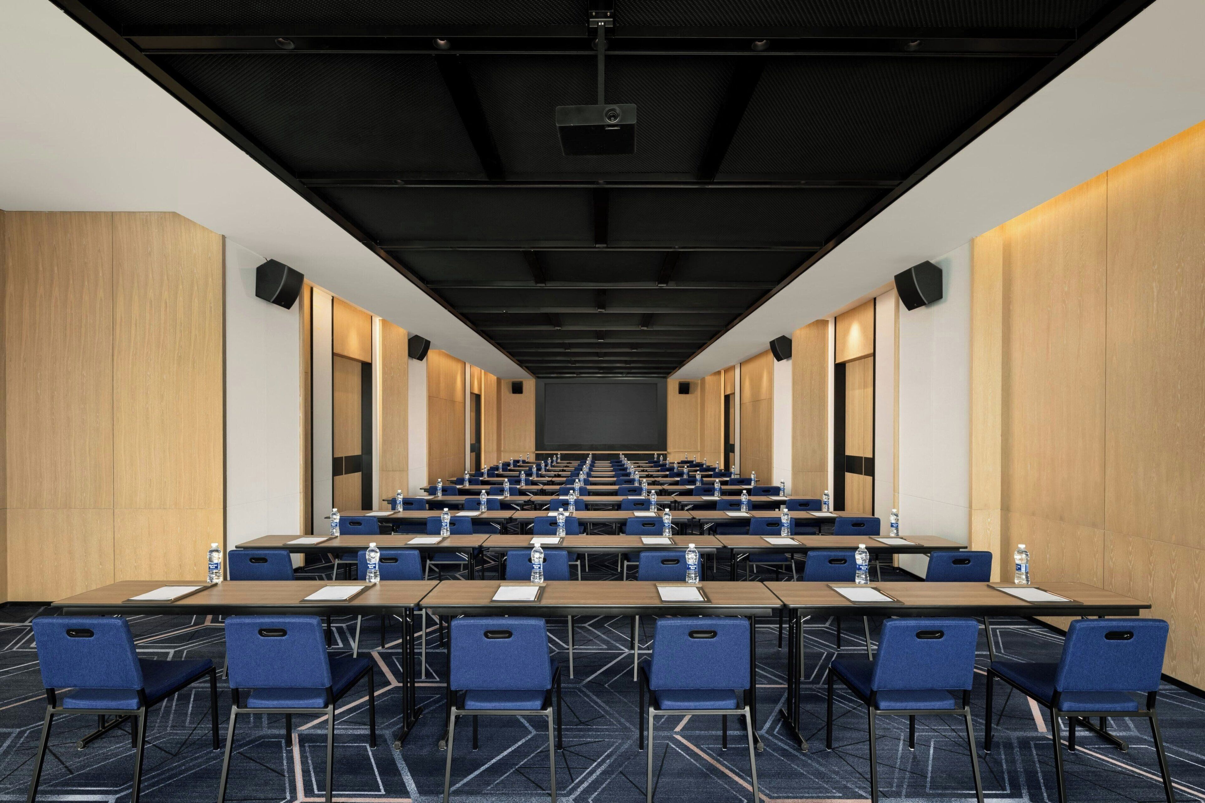 A conference room with rows of tables and chairs, a projector, and a large screen.