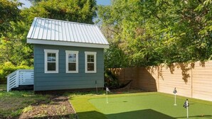 Une petite maison avec un toit en métal et des bordures blanches, située à côté d’un terrain synthétique vert, sur lequel sont plantés deux drapeaux de golf.