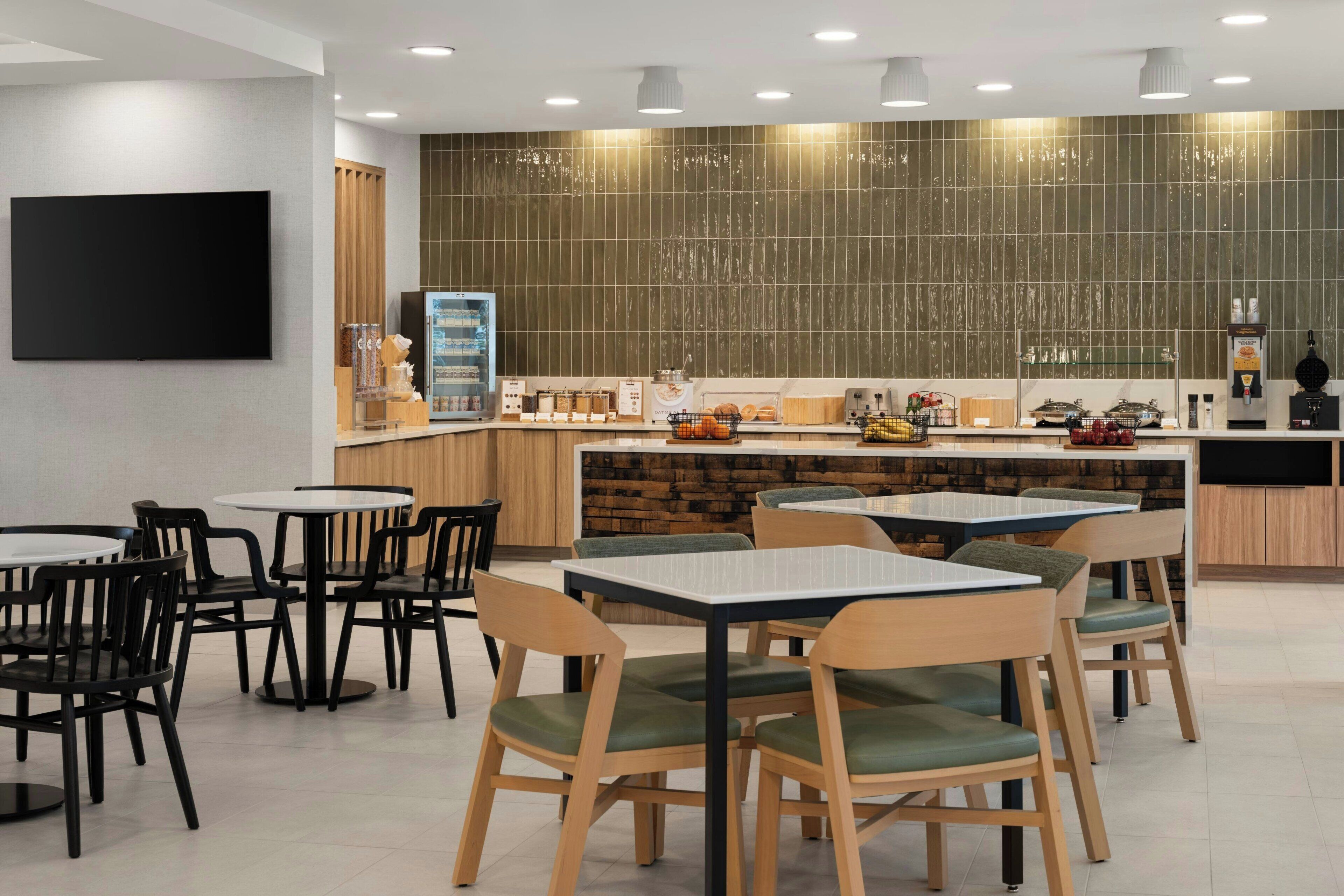 Een modern restaurant met ronde tafels, zwarte en houten stoelen, een flatscreen televisie en een keuken met een betegelde achterwand.