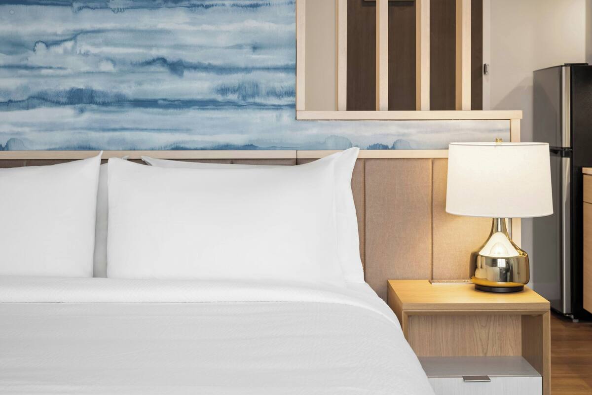 A hotel room with a bed, a nightstand with a lamp, a wooden headboard, a wall with a blue abstract design, and a refrigerator.