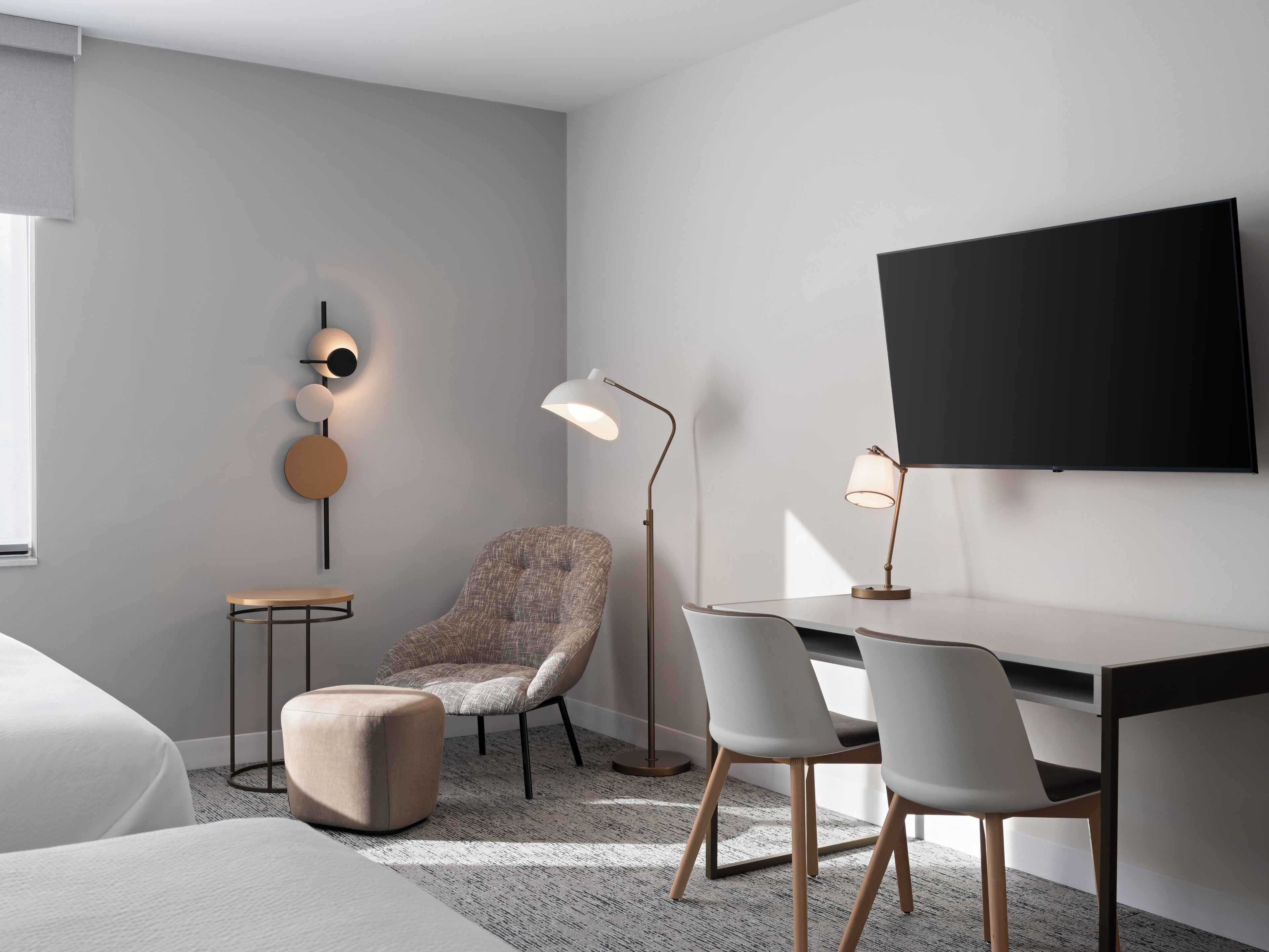 A modern hotel room with a bed, a desk with two chairs, a round table, a chair, a floor lamp, and a wall-mounted TV.