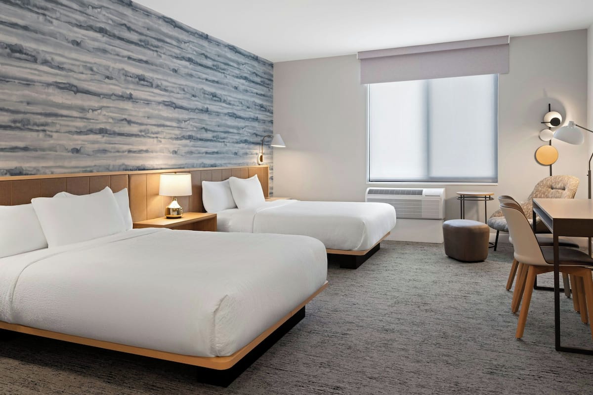 A hotel room with two beds, a desk, a chair, a lamp, and a large window.