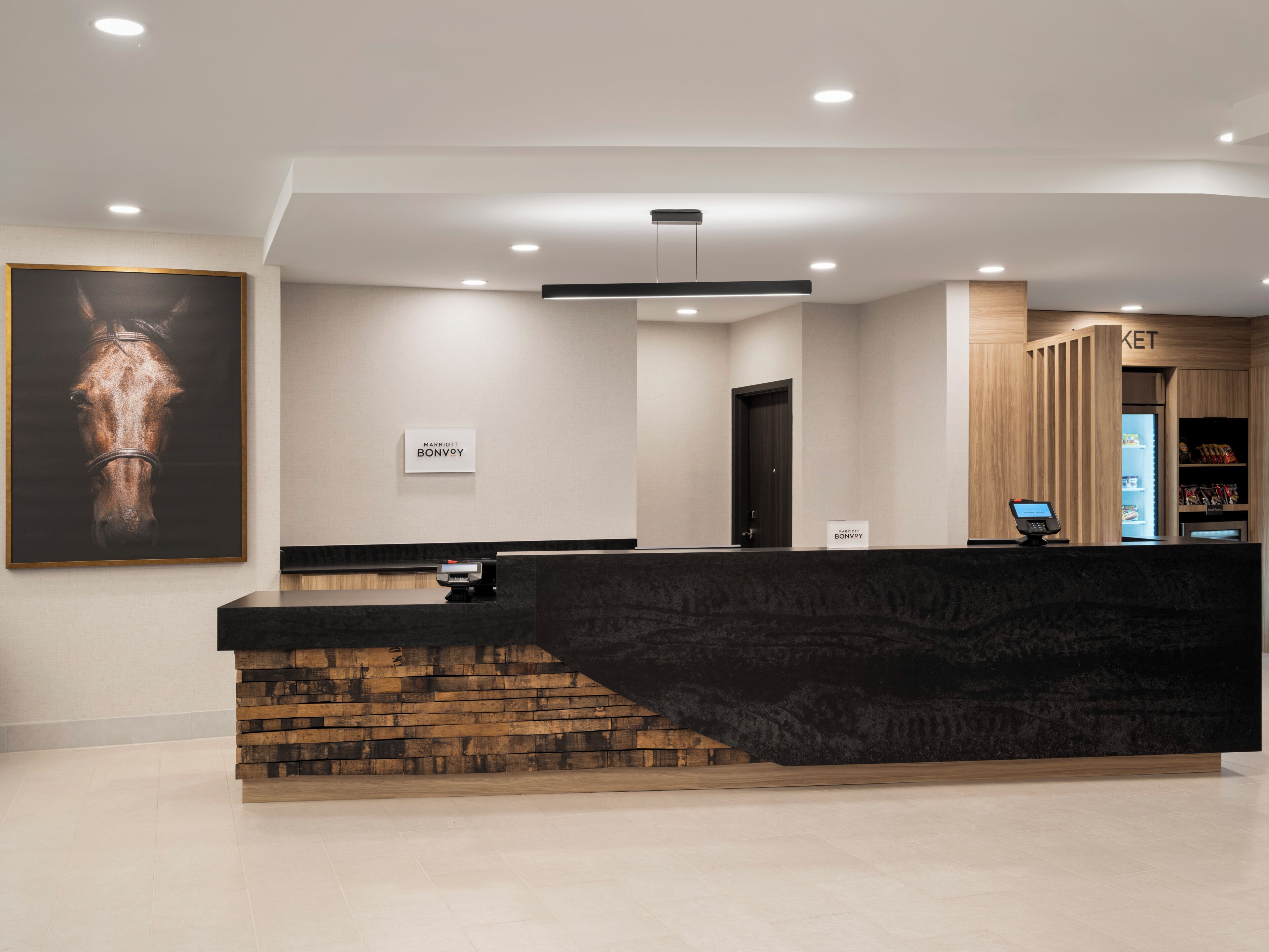 A hotel reception area with a modern design, a mounted horse portrait, a reception desk, a digital kiosk, and a minibar.