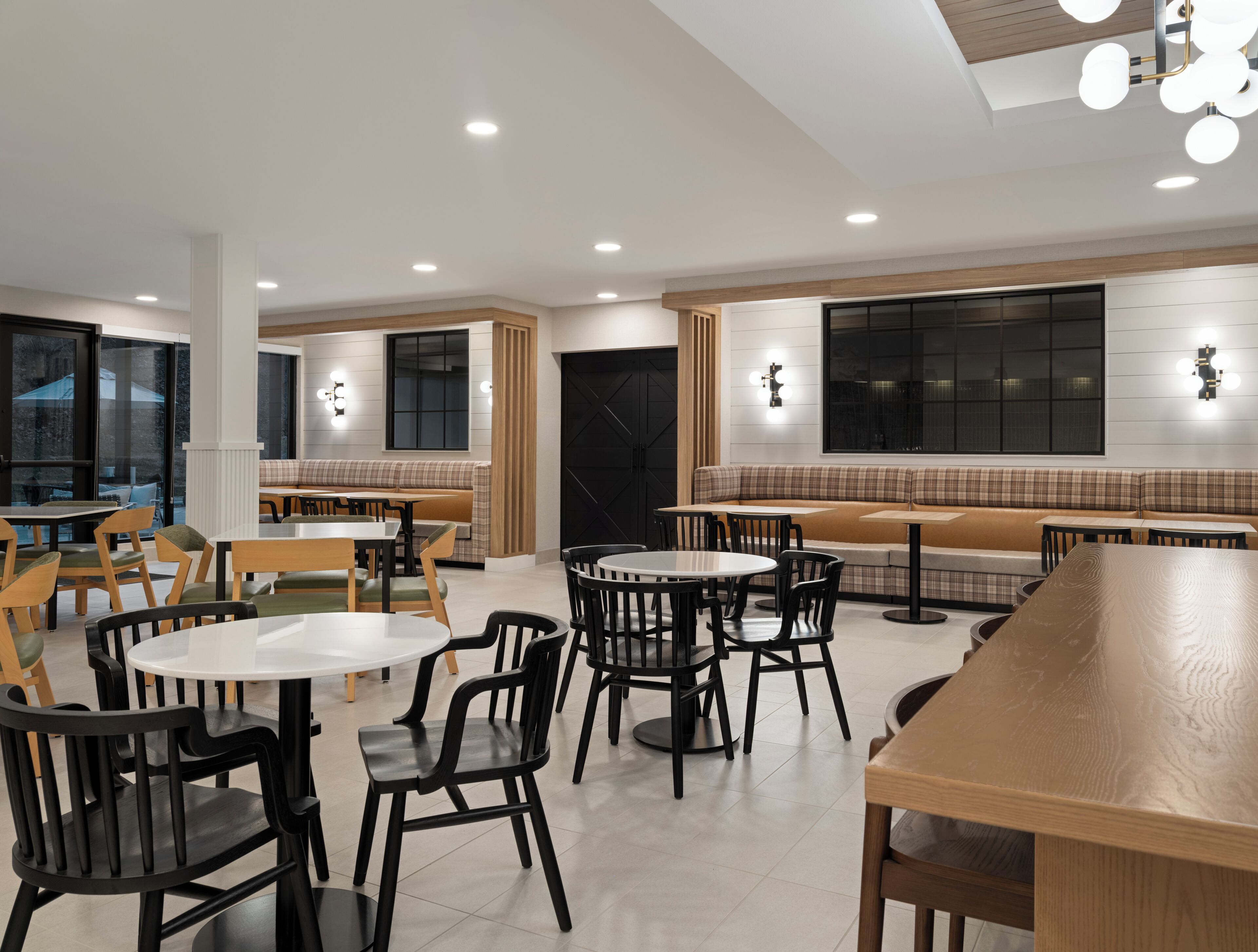 Interior moderno de restaurante com mesas redondas e cadeiras pretas, bancos de madeira e iluminação embutida no teto.