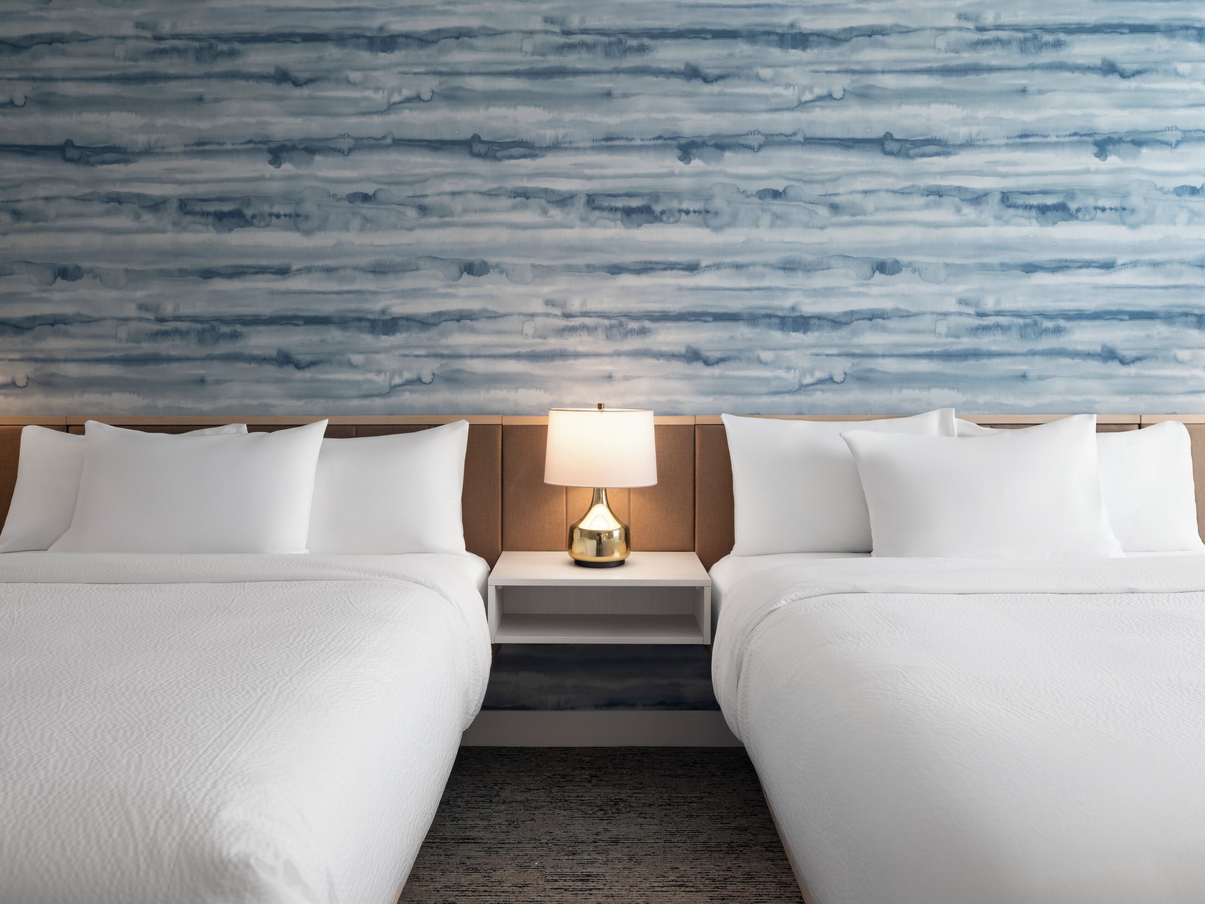 Camera d'albergo con due letti, un comodino con lampada e una parete azzurra con motivo astratto.